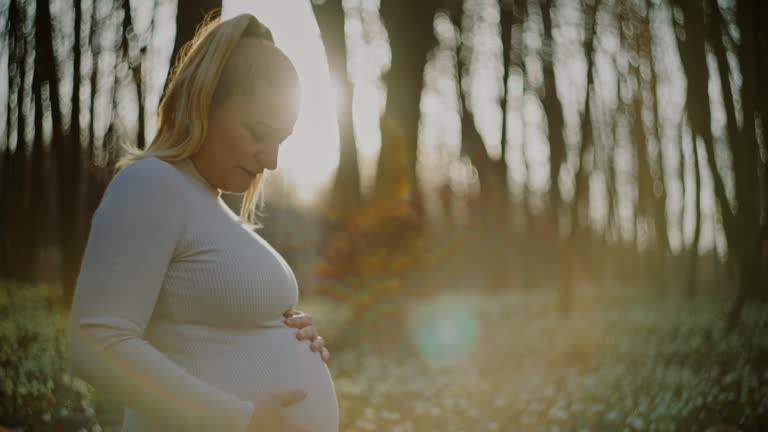 Portrait happy,serene pregnant woman holding baby bump among white snowdrop flowers in sunny,tranquil springtime woods. Shot in 8K Resolution.